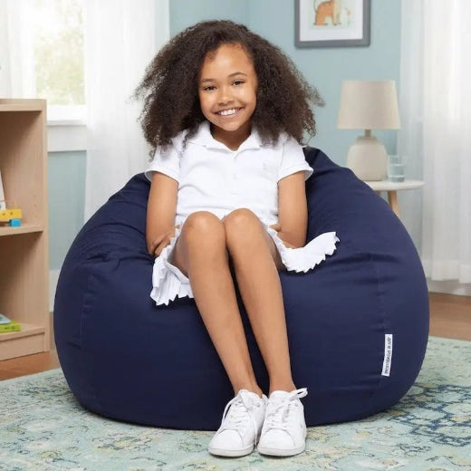 Young girl sitting on a navy bean bag chair in a room with a lamp and bookshelf.